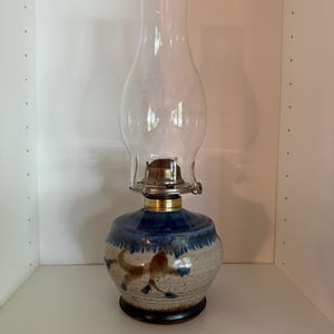May include: A ceramic oil lamp with a clear glass chimney. The base is a speckled beige with a blue band and brown floral design. The lamp has a brass collar and a black base.