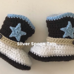 May include: A pair of handmade crochet baby cowboy boots. The boots are white and brown with a light blue star and trim. The boots have a tan strap with a button and a gray spur detail. The text "Silver Spoon Tots" is visible.