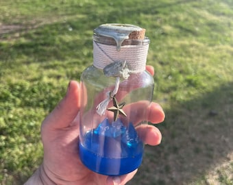 May include: A clear glass bottle with a cork stopper and blue liquid inside. The bottle is decorated with a star charm and white twine. The bottle is held in a person's hand.
