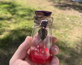 May include: A small glass bottle with a cork stopper, twine, and a decorative leaf charm. The bottle contains red liquid and several red dice. The cork is painted purple. The bottle is held in a hand.