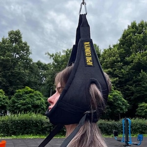 May include: A black neck traction device with the text "APNI DUNIYA" in yellow. The device is suspended from a metal carabiner. The person is wearing the device outdoors, with trees and exercise equipment in the background.
