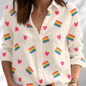May include: A white long-sleeve button-down shirt featuring a repeating pattern of small pink hearts and rectangular rainbow-colored blocks. The shirt has a classic collar, a front button placket, and long sleeves with cuffs, providing a clean look.