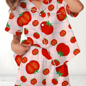 May include: Light pink pajama set with a tomato pattern. The short-sleeved top and shorts feature a grid design and are adorned with large red tomato illustrations. White trim outlines the edges of the pajamas.