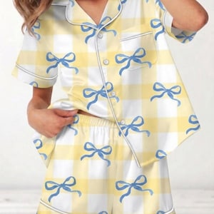 May include: Yellow and white gingham pajama set with blue bow accents. The short-sleeved top has a chest pocket and white piping. The matching shorts have a drawstring waist and white piping. The fabric has a satin sheen.