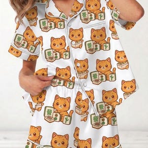 May include: White pajama set featuring an all-over pattern of orange cartoon cats playing mahjong. The short-sleeved top and shorts have white piping and buttons. The cats are depicted with various expressions and are surrounded by mahjong tiles and cacti.
