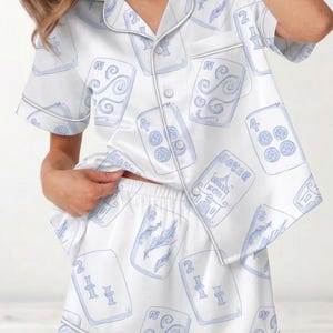 Puede incluir: Conjunto de pijama blanco con un estampado de cartas de juego en azul claro. La parte superior de manga corta y los pantalones cortos presentan un diseño repetido de palos de cartas, números y la palabra "Joker". El conjunto tiene ribetes y botones blancos.