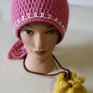 Può includere: Un cappello all'uncinetto fatto a mano di colore rosa polveroso con un bordo decorativo bianco. Sono attaccati una sciarpa abbinata e un piccolo sacchetto con coulisse giallo senape. Il cappello è esposto su una testa di manichino.