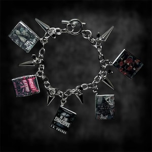 Op de afbeelding: Een zilveren kettingarmband met boekbedels en puntige accenten. De boekbedels tonen titels als "Her Soul to Take" en "In the Shadows". De armband heeft een tuimelsluiting.