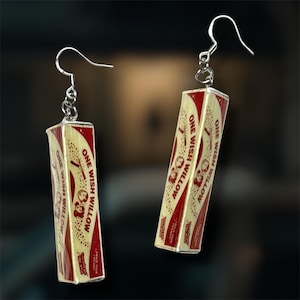 May include: A pair of dangle earrings featuring rectangular charms designed to resemble vintage gum packaging. The charms are cream-colored with red accents and text that reads ONE WISH WILLOW, attached to silver-toned metal earring hooks.