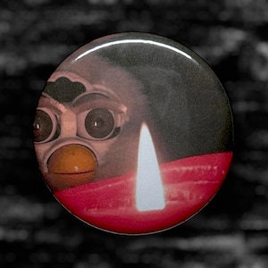Op de afbeelding: Ronde knoop met een close-up van een Furby-speelgoed met grote ogen en een gele snavel. Een witte kaarsvlam tegen een rode achtergrond, met een donkergrijze achtergrond.