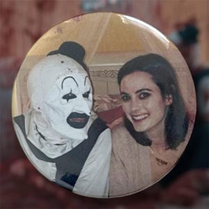 May include: Round button featuring a smiling woman and a person in a white clown mask with black accents. The button has a vintage, slightly faded appearance, with a soft, warm color palette.
