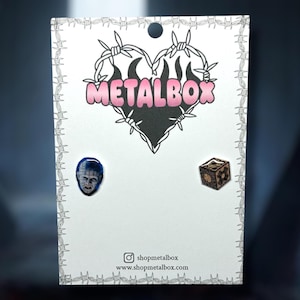 May include: A pair of stud earrings on a white card with a barbed wire border. One earring features a blue image of a person's face, and the other is a small wooden cube. The card has the text "METALBOX" in pink and the website address.