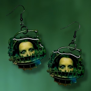 Könnte beinhalten: Ein Paar ausgefallene Ohrringe mit dem Gesicht einer Frau, das von einem grünen und metallfarbenen Kopfkäfig umrahmt wird. Die Ohrringe haben schwarze Metallhaken und ein gruseliges, Horror-Thema.