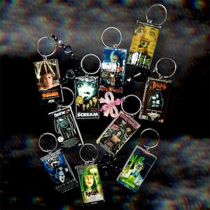 Pode incluir: Uma coleção de porta-chaves retangulares com cartazes de filmes de terror em miniatura, incluindo Nightmare on Elm Street, Scream, Dracula, The Lost Boys, Popcorn, Re-Animator e Frankenstein, cada um preso a um anel de metal prateado.