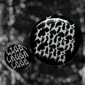 Puede incluir: Dos tapones para los oídos en blanco y negro con un patrón dentado repetitivo. Los tapones son circulares y tienen un acabado brillante. El fondo está borroso, con un efecto bokeh.
