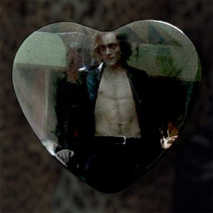 May include: Heart-shaped decorative item featuring a man in a black leather jacket and pants. The image is a close-up, with the man's torso visible. The background is blurred, with a dark, textured appearance.