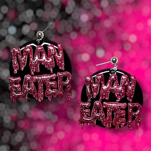 Può includere: Un paio di orecchini a lobo circolari con base nera e la scritta MAN EATER in un carattere rosa scuro glitterato e gocciolante. Gli orecchini sono fissati a perni e chiusure in metallo color argento, con un design audace e contrastato.