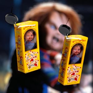 Puede incluir: Pendientes con cajas amarillas en miniatura que muestran una muñeca y el texto "Good Guys". Las cajas tienen ilustraciones de naipes. Cada pendiente tiene una parte superior circular negra.