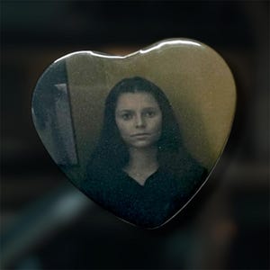 May include: A heart-shaped pin featuring a glossy finish and a centered photographic portrait of a person with long dark hair. The image has a muted, sepia-toned aesthetic with a dark background, and the pin has a smooth, reflective surface.