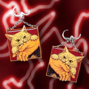 May include: Two cat-themed earrings. Each earring features a square design with a cartoon cat biting a hand. The cat is yellow with yellow eyes, and the hand is orange. The background is red. The earrings have silver-colored hooks and chains.