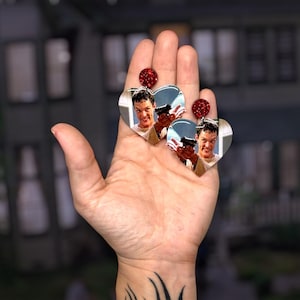 Könnte beinhalten: Herzförmige Ohrringe mit einem Mann, der eine Waffe hält, und einem roten Glitzerdetail. Die Ohrringe werden vor einem verschwommenen Hintergrund präsentiert. Ein ausgefallenes Accessoire.