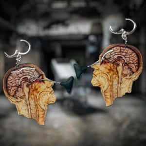 Puede incluir: Un par de pendientes únicos con un diseño de sección transversal de cabeza humana. Los pendientes son de madera y tienen un gancho plateado. El diseño incluye un cerebro y una forma triangular negra.