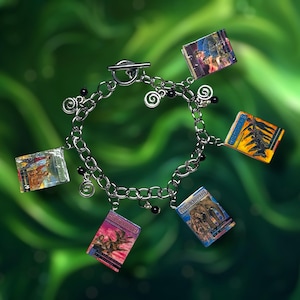 Könnte beinhalten: Ein silbernes Charm-Armband mit sechs kleinen, rechteckigen Anhängern. Jeder Anhänger zeigt eine farbenfrohe Illustration, die wie Miniaturbücher aussehen. Das Armband hat einen Knebelverschluss und ist vor einem verschwommenen grünen Hintergrund platziert.