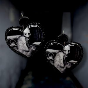 May include: Black heart-shaped earrings with a glittery border. Each earring features a black and white image of a pale, gaunt figure. The earrings have a post-back closure and are designed for pierced ears.