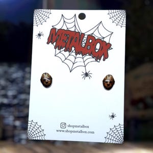 Op de afbeelding: Oorknopjes met een bruin en wit hockeymasker ontwerp. Ze worden gepresenteerd op een witte kaart met een rood "METALBOX" logo, spinnenwebben en spinnen. Social media informatie is ook zichtbaar op de kaart.