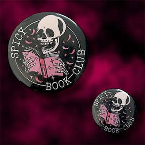 May include: Two black round buttons with a skull reading a pink book, surrounded by stars and moons. The text "SPICY BOOK CLUB" is printed around the edge in white. The buttons are set against a dark, textured background.