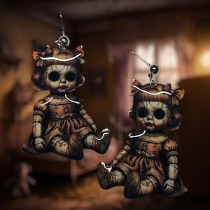 Könnte beinhalten: Ein Paar gruselige Puppenohrringe. Jeder Ohrring zeigt eine sitzende Puppe mit einem verwitterten, Vintage-Look. Die Puppen haben dunkle Augen, genähte Merkmale und tragen braune Kleider mit Schleifen. Die Ohrringe hängen an silberfarbenen Haken.