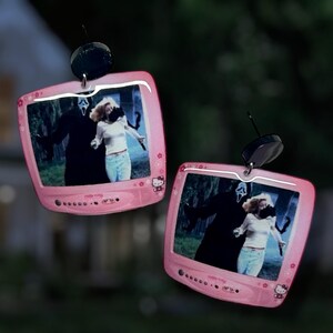 Könnte beinhalten: Ein Paar rosa Ohrringe in Form eines Retro-Fernsehers. Jeder Ohrring zeigt eine Szene aus dem Film "Scream" und ist mit Hello Kitty-Details verziert. Die Ohrringe haben schwarze, kreisförmige Aufsätze oben.
