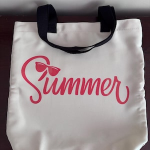 May include: Cream canvas tote bag with black handles. The word "Summer" is printed in red script with sunglasses incorporated into the design. The bag is designed for carrying items.
