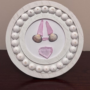 May include: A round, white wooden frame with a beaded border. Inside, a design features pink sea glass and seashells arranged on a white background. The arrangement resembles a face. A unique piece of coastal decor.
