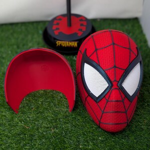Pu&ograve; includere: Una maschera rossa di Spider-Man con ragnatele nere e lenti bianche per gli occhi. Sono visibili anche un guscio facciale rosso e un supporto nero con la scritta "SPIDERMAN". Gli oggetti sono su una superficie verde.