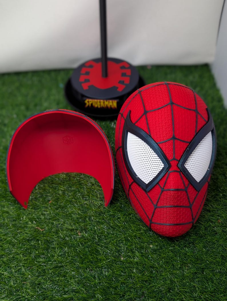 Pu&ograve; includere: Una maschera rossa di Spider-Man con ragnatele nere e lenti bianche per gli occhi. Sono visibili anche un guscio facciale rosso e un supporto nero con la scritta "SPIDERMAN". Gli oggetti sono su una superficie verde.