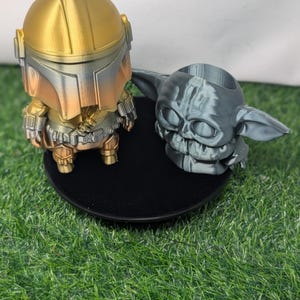 May include: Two 3D-printed figurines: a Mandalorian character in gold, silver, and bronze tones, and a gray Grogu figure. The Mandalorian stands on a black base. The Grogu figure has a hollowed-out head, possibly for use as a planter.