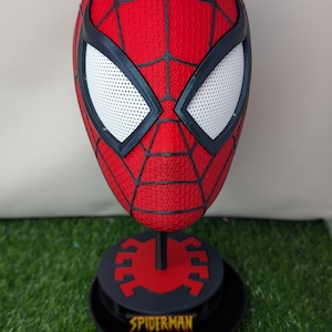 Pu&ograve; includere: Una maschera rossa di Spider-Man con ragnatele nere e lenti bianche. La maschera &egrave; esposta su un supporto nero con un emblema di ragno rosso e la scritta "SPIDERMAN" in giallo.