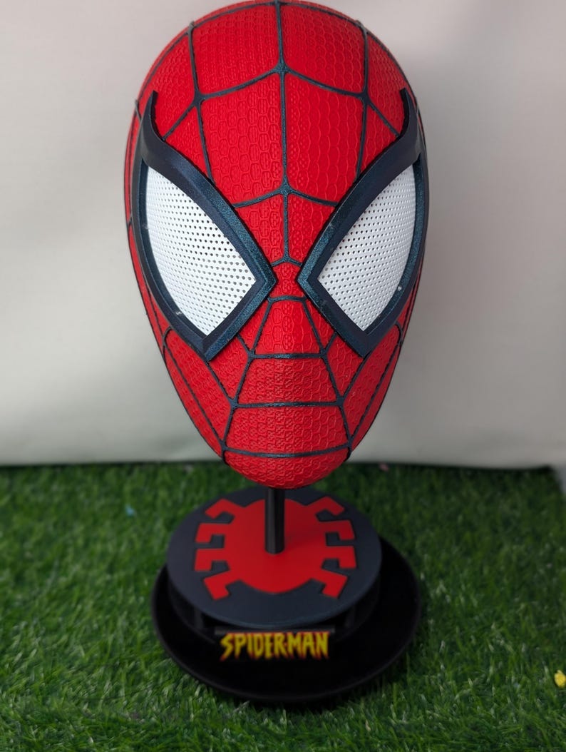 Pu&ograve; includere: Una maschera rossa di Spider-Man con ragnatele nere e lenti bianche. La maschera &egrave; esposta su un supporto nero con un emblema di ragno rosso e la scritta "SPIDERMAN" in giallo.