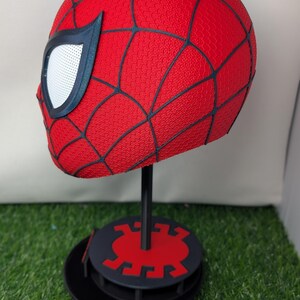 Pu&ograve; includere: Un casco rosso di Spider-Man con linee nere a ragnatela e lenti bianche per gli occhi. Il casco &egrave; esposto su un supporto nero con una base rossa e nera, su sfondo verde.