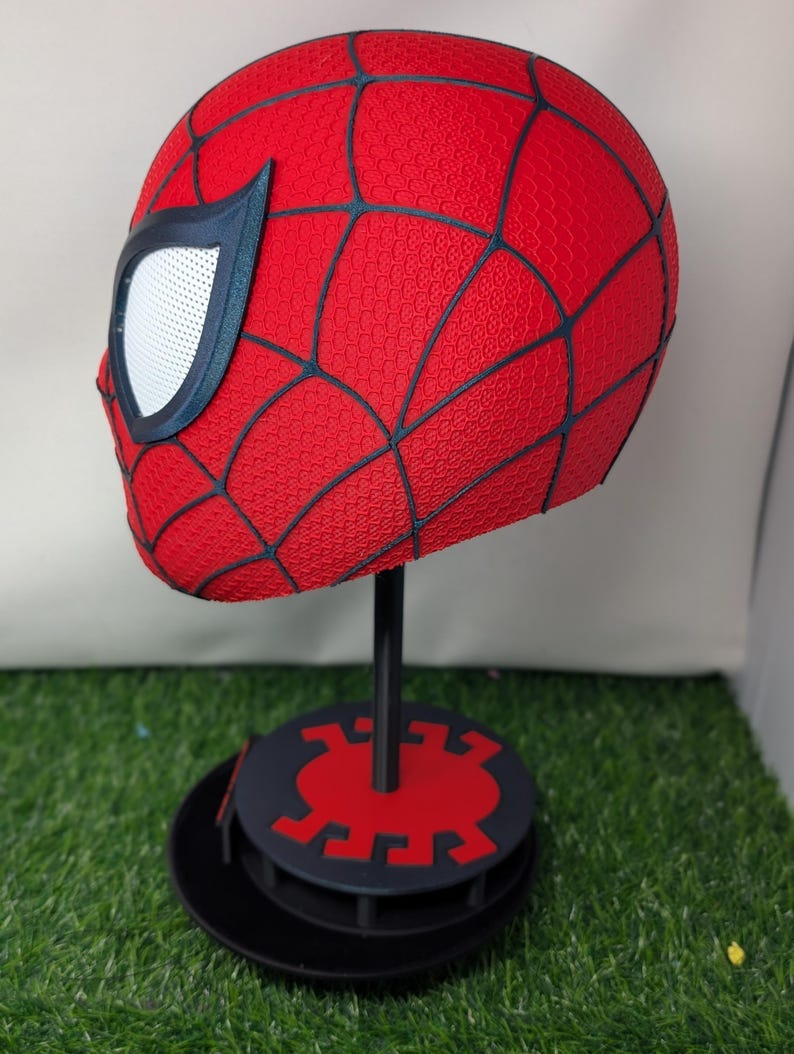 Pu&ograve; includere: Un casco rosso di Spider-Man con linee nere a ragnatela e lenti bianche per gli occhi. Il casco &egrave; esposto su un supporto nero con una base rossa e nera, su sfondo verde.