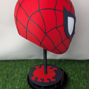 Pu&ograve; includere: Una maschera rossa di Spider-Man con dettagli neri a ragnatela e lenti bianche per gli occhi. La maschera &egrave; esposta su un supporto nero con base rossa e nera, su sfondo verde.