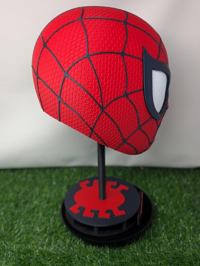 Pu&ograve; includere: Una maschera rossa di Spider-Man con dettagli neri a ragnatela e lenti bianche per gli occhi. La maschera &egrave; esposta su un supporto nero con base rossa e nera, su sfondo verde.