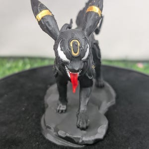 May include: A black and gold 3D-printed figure of a creature with a red tongue. The figure has large ears with gold accents and a white and gold eye design. It stands on a gray, textured base.