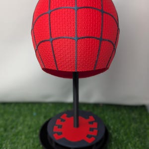Pu&ograve; includere: Un oggetto sferico rosso con una superficie strutturata e linee a griglia nere, montato su un supporto nero con una base circolare. La base presenta un emblema di ragno rosso. L'articolo &egrave; esposto su una superficie verde.