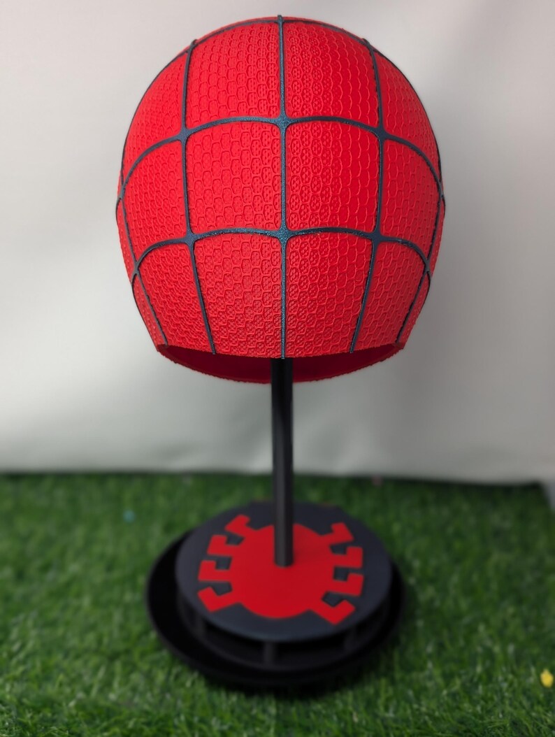 Pu&ograve; includere: Un oggetto sferico rosso con una superficie strutturata e linee a griglia nere, montato su un supporto nero con una base circolare. La base presenta un emblema di ragno rosso. L'articolo &egrave; esposto su una superficie verde.