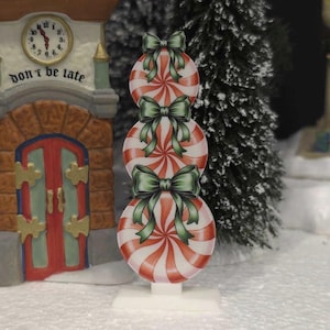 May include: A decorative Christmas village scene featuring a stack of three red and white peppermint candy ornaments with green bows. The scene includes miniature buildings, a clock tower with the text "don't be late," and a snowy landscape.