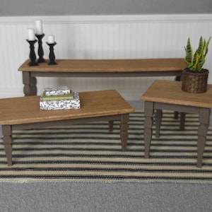 May include: Three wooden coffee tables in a living room setting. The largest table is a rectangular coffee table with a brown wood top and black legs. The middle table is a rectangular coffee table with a brown wood top and brown legs. The smallest table is a square end table with a brown wood top and brown legs. A green plant in a brown pot sits on the end table. A striped rug is on the floor.