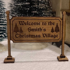 May include: A wooden sign with the text "Welcome to the Smith's Christmas Village" is displayed. The sign is brown with a decorative border and two Christmas trees. The sign is supported by two posts and is part of a Christmas village display.