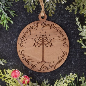 May include: A round wooden Christmas ornament featuring a tree design with text around the edge. The ornament is surrounded by green and white holiday foliage with red berries. A jute string is attached for hanging.